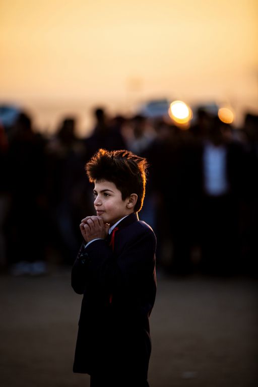 Ahmed, age 8 and cousin of the groom, looks on as Amera and Samir dance as the sun sets following their wedding ceremony on December 29, 2018. The celebration took place in a small clearing outside of the Bajed Kandala Yazidi Displacement Camp in Iraqi Kurdistan; all those in attendance are displaced Yazidis and returned to their tents within the fenced camp following the celebration.This image was taken on December 29, 2018.